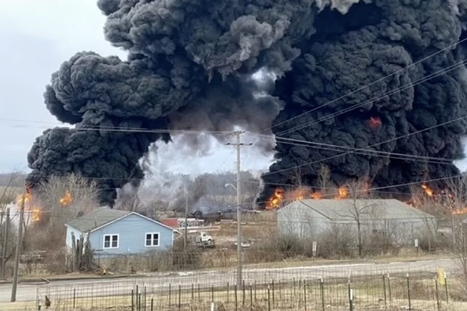 A plume of black smoke behind a small house in a rural setting.